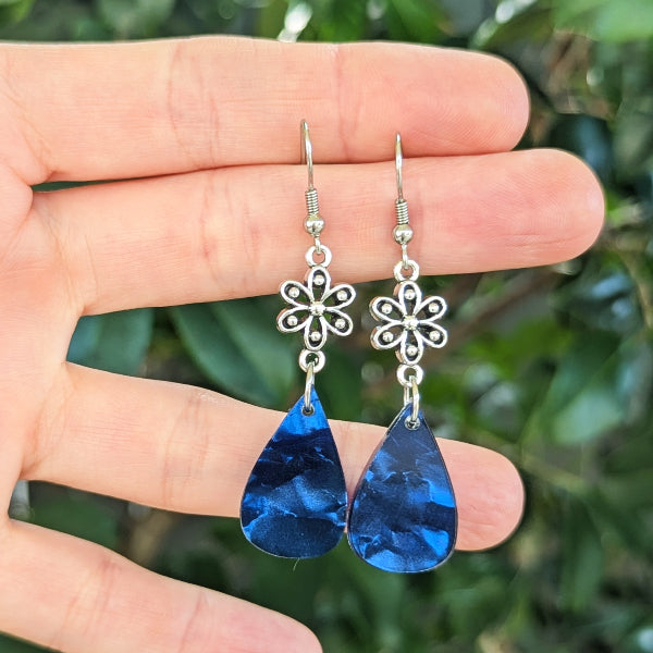 A pair of dangling earrings held between fingers, featuring silver-tone filigree flower charms and deep blue teardrop-shaped drops, with a blurred green outdoor background.