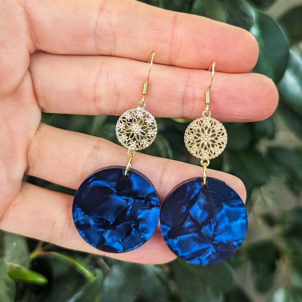 A pair of dangling earrings held in a hand, featuring gold-tone hook wires, circular filigree disc charms, and large marbled deep blue disc drops, with a blurred green leafy background.