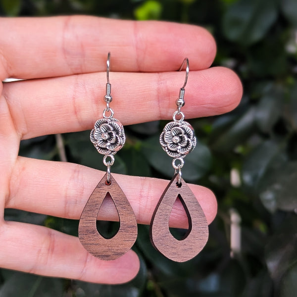 A pair of handmade dangle earrings held in a hand, featuring silver-toned metal flower charms and teardrop-shaped wooden pendants with a natural brown wood grain, set against a blurred background of green leaves.