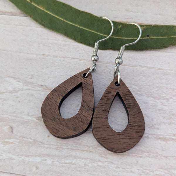A pair of teardrop-shaped wooden drop earrings with a cutout center and silver fishhook wires, displayed on a light wood surface with a green leaf in the background.