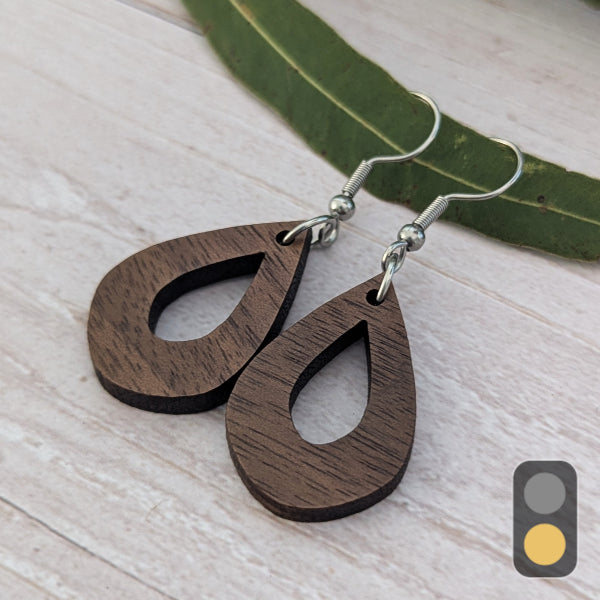 Teardrop-shaped wooden drop earrings with a dark brown wood grain finish and silver fishhook ear wires, displayed on a light wood surface next to a green leaf.