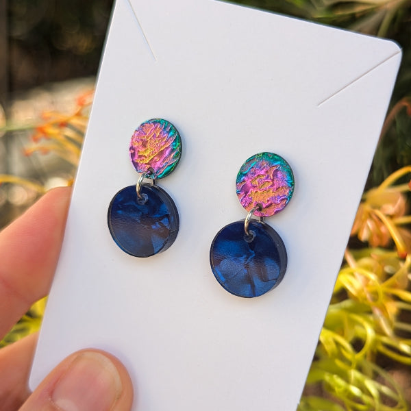 A pair of handmade dangle earrings displayed on a white card, held by a hand against a blurred outdoor background of green foliage and warm light. The earrings feature a top disc with a colorful, textured metallic finish in pink, purple, and green, hanging from a silver ring from a larger, dark blue circular drop.