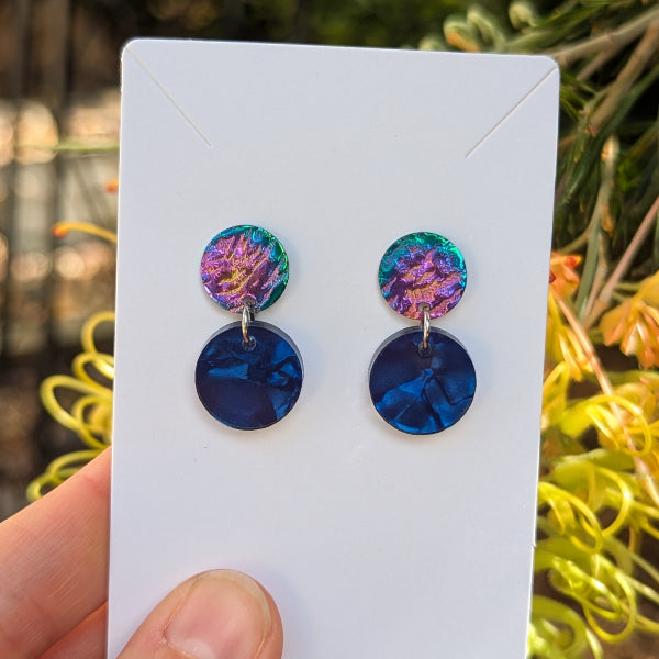 A pair of drop earrings displayed on a white card, held by a hand against a blurred outdoor background of green foliage and yellow flowers. The earrings feature a top disc with a purple and green iridescent pattern and a bottom disc in deep blue.