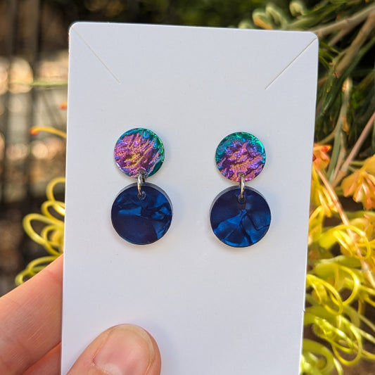 A pair of drop earrings displayed on a white card, held by a hand against a blurred outdoor background of green foliage and yellow flowers. The earrings feature a top disc with a purple and green iridescent pattern and a bottom disc in deep blue.