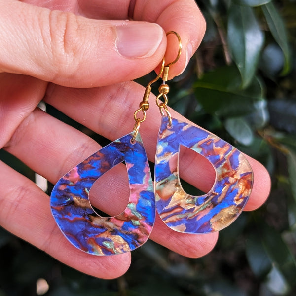 A pair of teardrop-shaped drop earrings with gold look hook wires and iridescent blue, pink, and gold marbled resin centers, held in a person's hand against a blurred green leaf background.