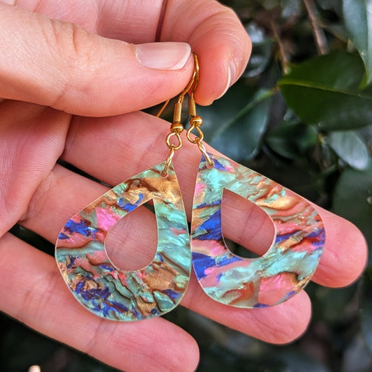 A pair of teardrop-shaped dangling earrings with a marbled iridescent finish in teal, gold, pink, and blue, held in a hand against a blurred background of green leaves.