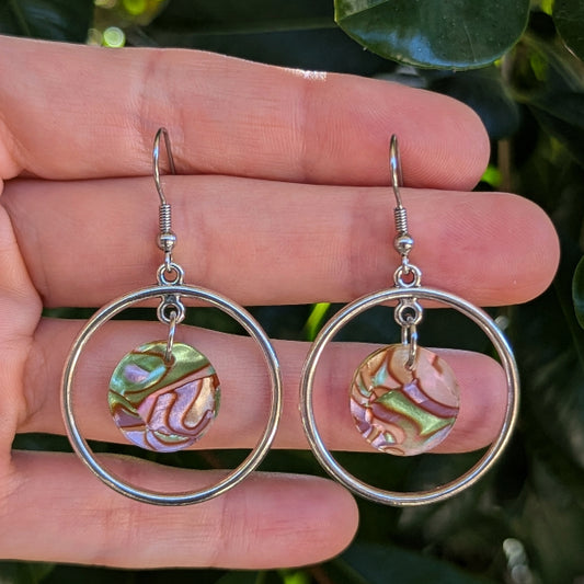A pair of silver-tone hoop earrings with round iridescent acrylic disc charms, held in a person's hand against a background of green leaves.