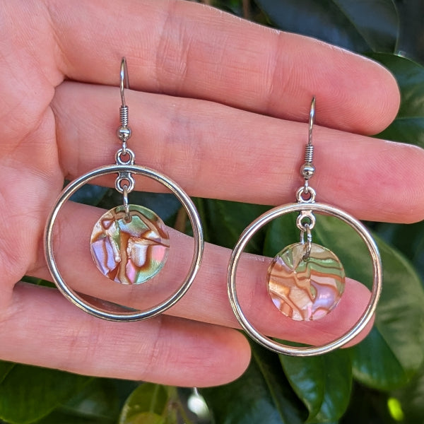 A pair of silver-toned dangle earrings with circular hoops and round iridescent acrylic pendants, held in a person's hand against a background of green leaves.
