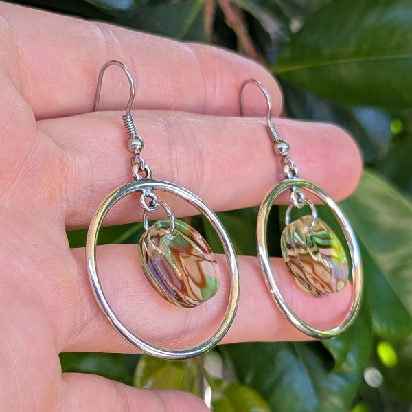 A pair of silver-tone hoop earrings with oval drop hoops and marbled olive green, brown, and gold acrylic discs, held in a hand against a background of green leaves.