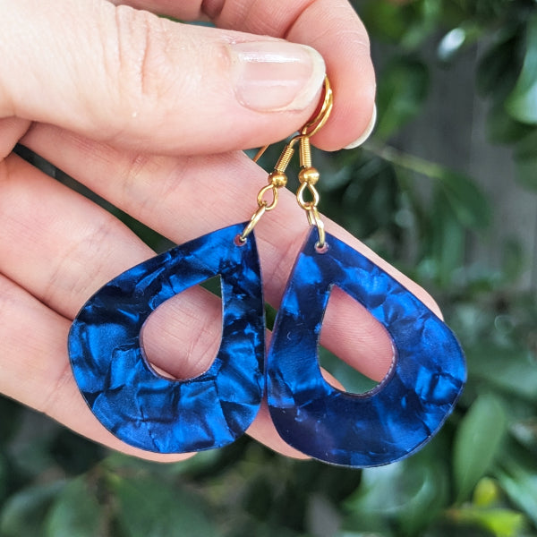 A close-up of a hand holding a pair of blue marbled teardrop-shaped dangle earrings with gold hook ear wires against a blurred green outdoor background.