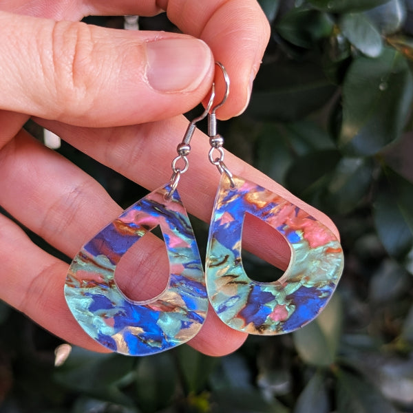 Teardrop-shaped dangling earrings with a marbled iridescent pattern in blue, green, pink, and gold, held by a hand against a background of dark green leaves.