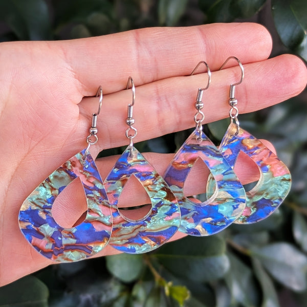 Two pairs of teardrop-shaped dangling earrings with a marbled blue, green, and pink acrylic pattern, held on a person's fingers against a background of green leaves.