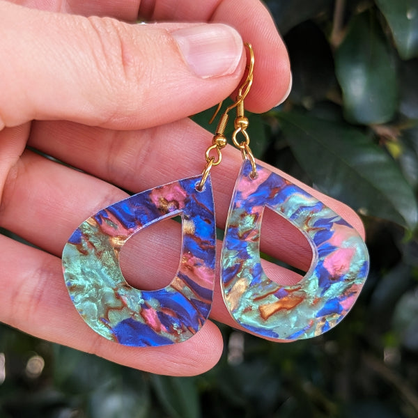 A close-up of a hand holding a pair of teardrop-shaped earrings with gold fishhook wires and iridescent abalone acrylic  inlays showing blue, pink, and green tones, set against a blurred background of green leaves.