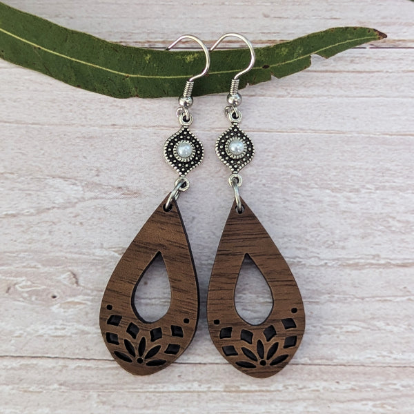 A pair of bohemian-style wooden dangle earrings with laser-cut floral patterns, featuring silver-tone hooks and bezel-set pearl-like accents, displayed on a green leaf over a whitewashed wooden background.