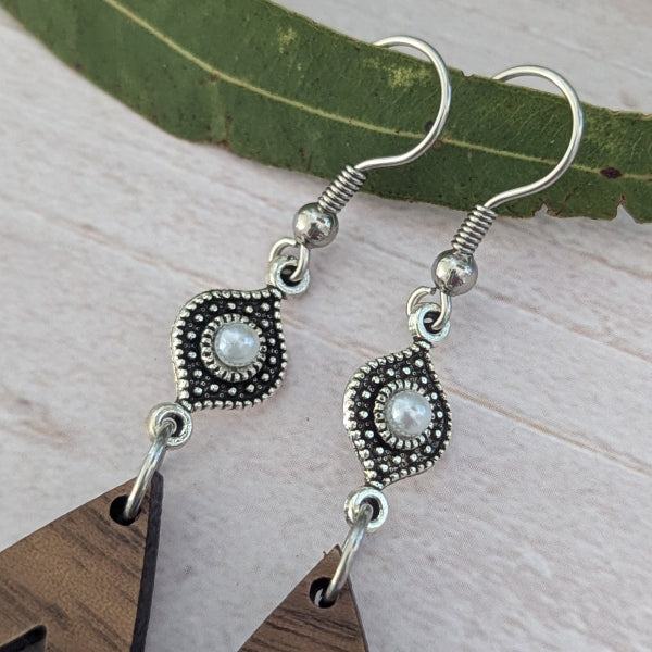A pair of silver-toned dangle earrings with ornate filigree details and oval white pearl-like centers, shown hanging against a light wood surface with a green leaf behind them.