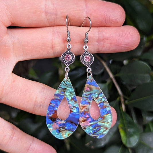 A pair of teardrop-shaped dangle earrings with iridescent blue, green, and purple acrylic pieces and silver hooks, held in a person's hand against a background of green leaves.