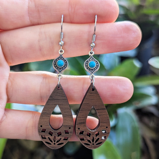 Wooden teardrop earrings with turquoise stones held between fingers against a blurred green background. Stainless steel hooks.