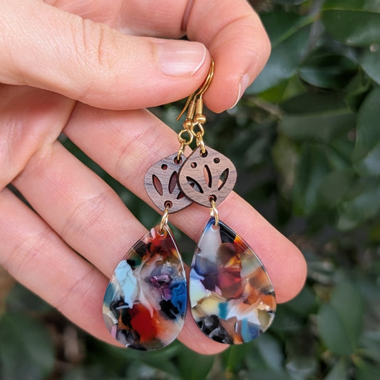 Hand holding a pair of earrings with colorful teardrop-shaped pendants against a green leafy background.