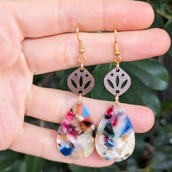Colorful teardrop earrings held between fingers with a green leafy background