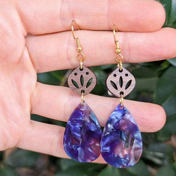 Purple teardrop earrings with gold hooks held in a hand against a blurred green leafy background