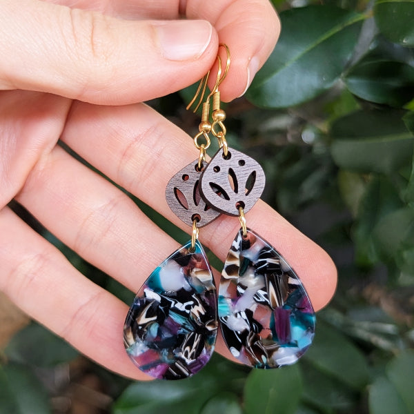 Hand holding a pair of earrings with floral designs against a green leafy background