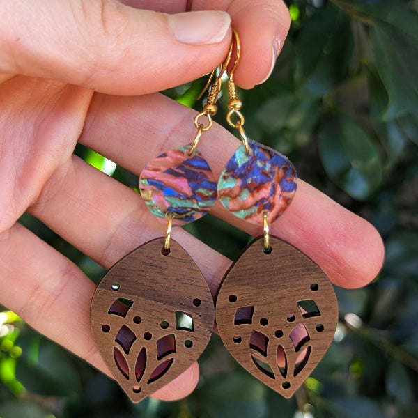 A pair of handmade dangle earrings held in a hand, featuring iridescent acrylic discs with pink, blue, and green hues, and wooden leaf-shaped drops with laser-cut floral patterns.