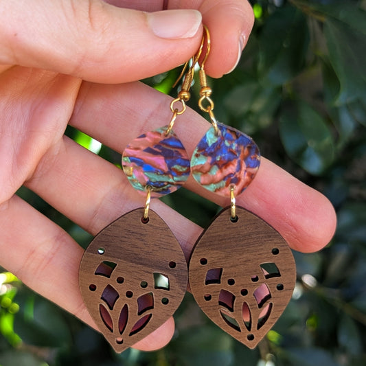 A pair of handmade dangle earrings held in a hand, featuring iridescent acrylic discs with pink, blue, and green hues, and wooden leaf-shaped drops with laser-cut floral patterns.