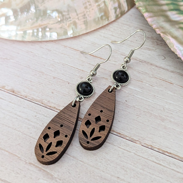 A pair of bohemian-style wooden dangle earrings with laser-cut floral patterns, black stones, and silver-tone hook wires, displayed on a light wood surface.