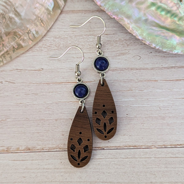 A pair of bohemian-style wooden dangle earrings with laser-cut floral patterns, dark blue oval stones, and silver-tone hook wires, displayed on a light wood surface.