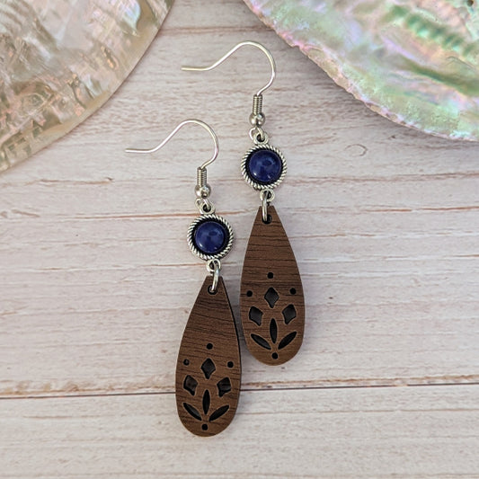 A pair of bohemian-style wooden dangle earrings with laser-cut floral patterns, dark blue oval stones, and silver-tone hook wires, displayed on a light wood surface.