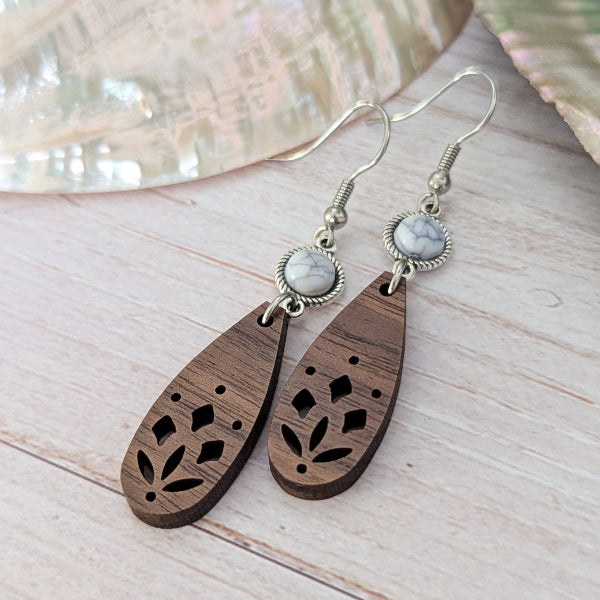 A pair of bohemian-style wooden dangle earrings with laser-cut floral patterns, white/grey stones, and silver-tone hook wires, displayed on a light wood surface.
