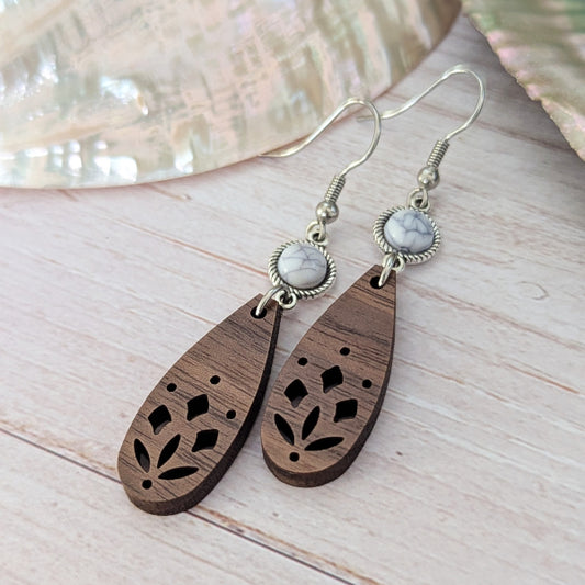 A pair of bohemian-style wooden dangle earrings with laser-cut floral patterns, white/grey stones, and silver-tone hook wires, displayed on a light wood surface.