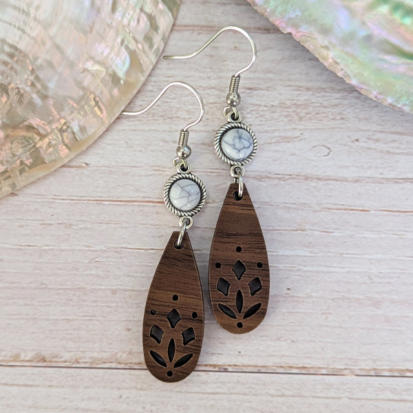 A pair of bohemian-style wooden dangle earrings with laser-cut floral patterns, white/grey stones, and silver-tone hook wires, displayed on a light wood surface.