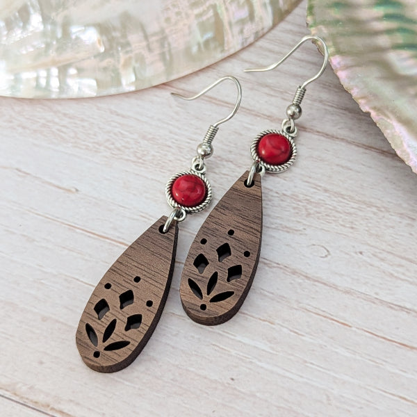A pair of bohemian-style wooden dangle earrings with laser-cut floral patterns, red stones, and silver-tone hook wires, displayed on a light wood surface.