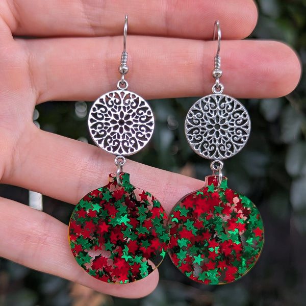 A pair of handmade dangle earrings held in a hand. The earrings feature silver-toned filigree disc tops and round green and red glitter drop charms.