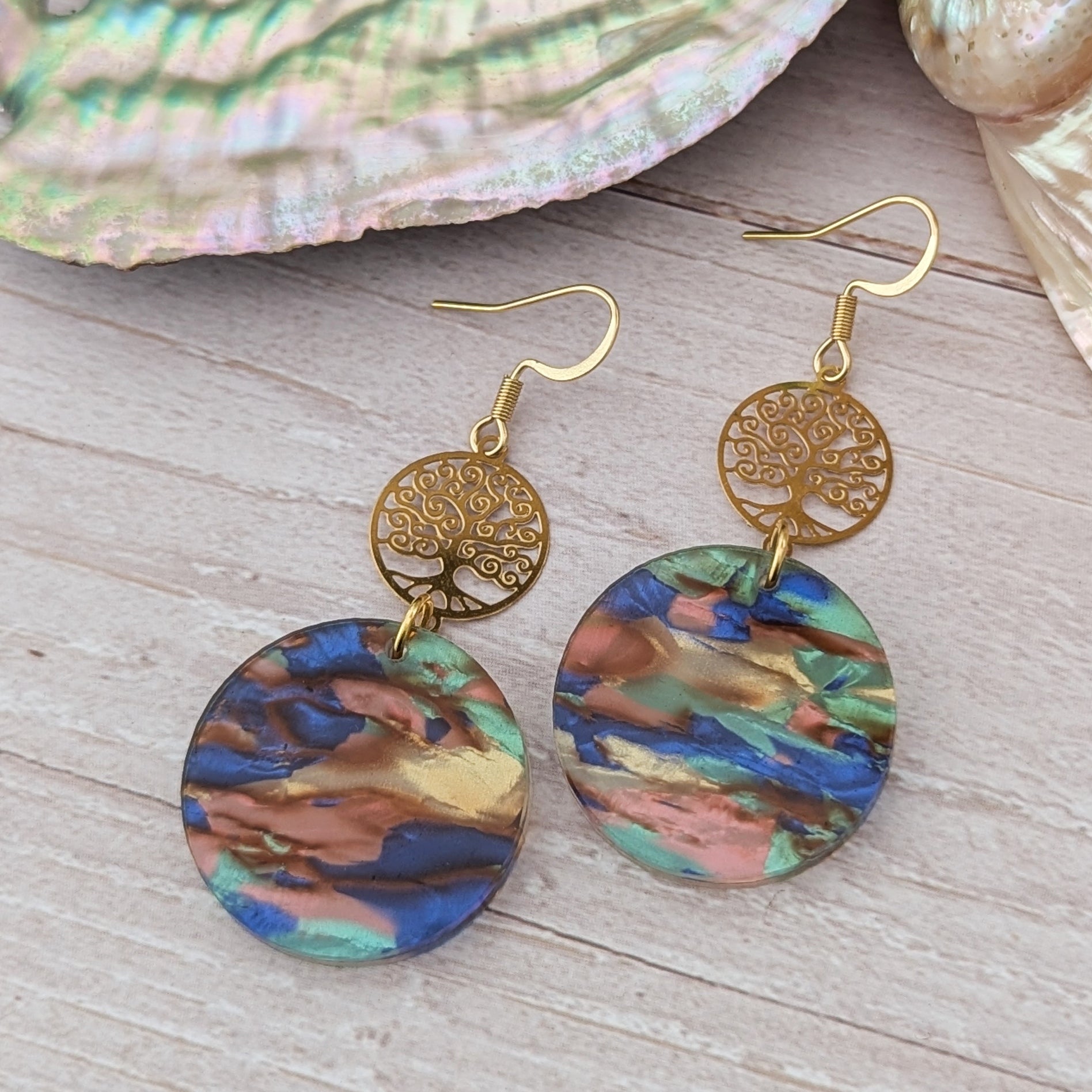 Colorful earrings with gold tree-like designs on a wooden surface with seashells.