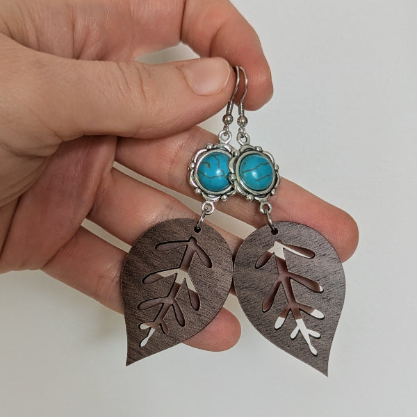 Hand holding a pair of earrings with wooden leaf design and turquoise stones.