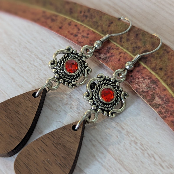 A pair of dangling earrings with ornate antiqued silver filigree, round red, and teardrop-shaped dark brown wooden drops, displayed on a light wood surface with a dried leaf.