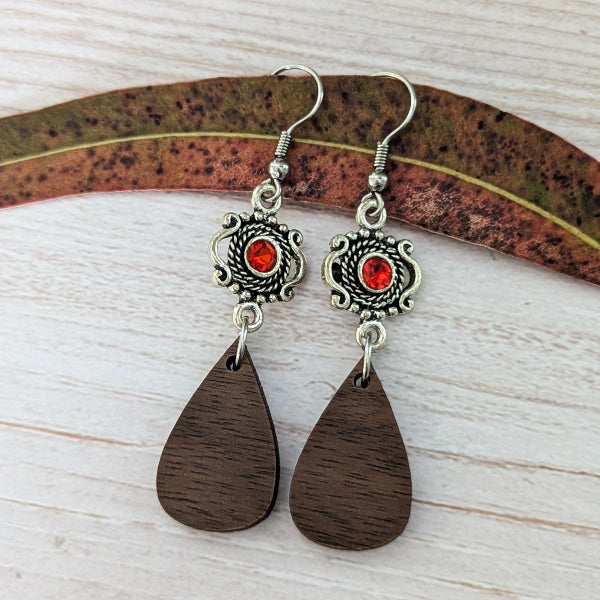 A pair of handmade dangle earrings with silver filigree frames, red centers, and dark brown wooden teardrop pendants, displayed on a leaf over a light wood background.