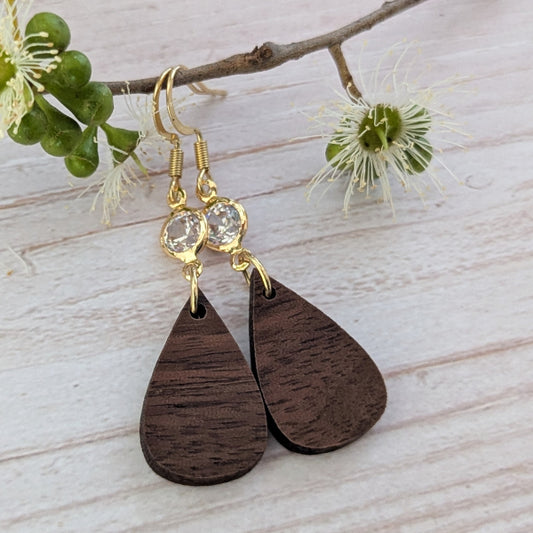 Wooden teardrop earrings with gold and crystal accents on a light wooden surface with flowers. Gold plated stainless steel hooks.