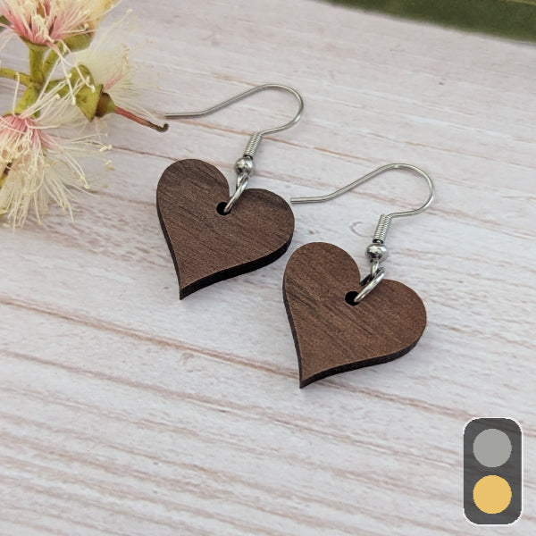 Heart-shaped wooden earrings on a light wooden surface with flowers in the background. Stainless steel hooks.
