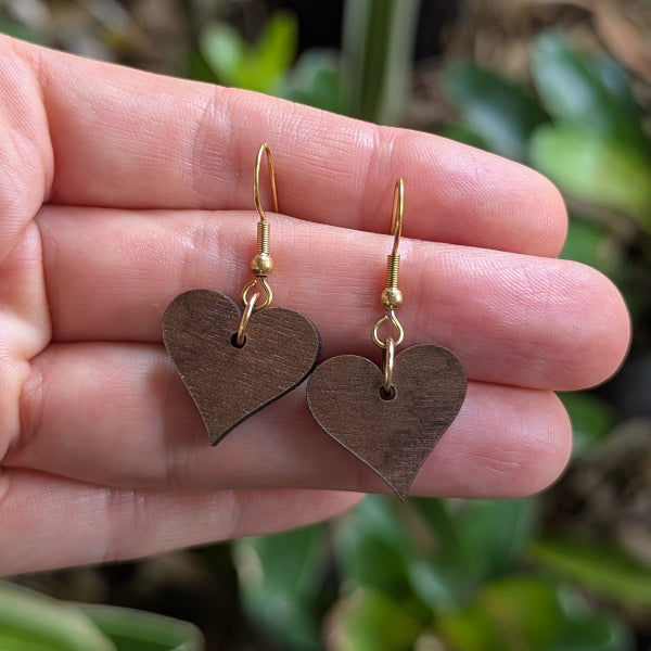 Heart-shaped earrings held in a hand with a green plant background. Gold plated stainless steel hooks.