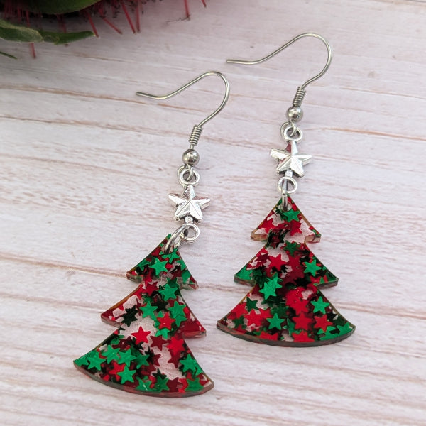A pair of dangle earrings shaped like Christmas trees, featuring red and green star-patterned resin pendants with silver-tone fishhook wires and star charms, displayed on a light wood background.