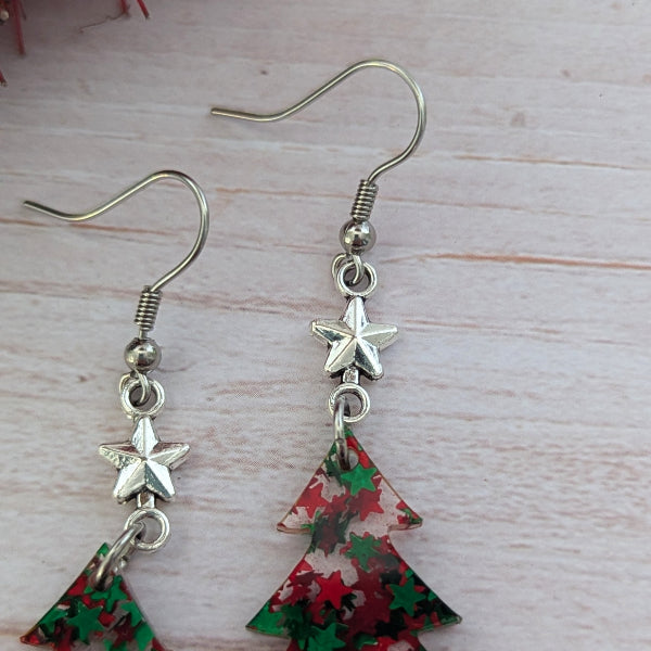 A pair of dangle earrings with silver-toned fishhook wires, star charms, and triangular Christmas tree-shaped charms decorated with a red and green holiday pattern, displayed on a light wood background.