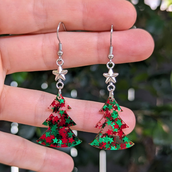 A pair of dangling earrings held between fingers. Each earring has a silver fishhook and star charm above a glittery Christmas tree shape in red and green.