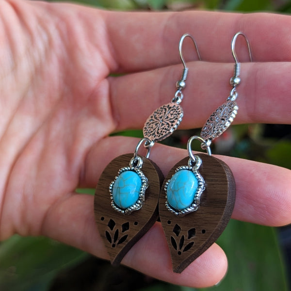 leaf-shaped earrings with turquoise stones and stainless steel hooks held between fingers against a blurred natural background.