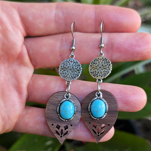 Pair of wooden earrings with turquoise stones held between fingers against a blurred natural background