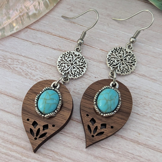 Wooden teardrop earrings with turquoise stones and silver floral designs on a light wooden surface.