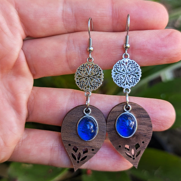 Pair of earrings with wooden leaf design and blue stones held between fingers against a natural background.