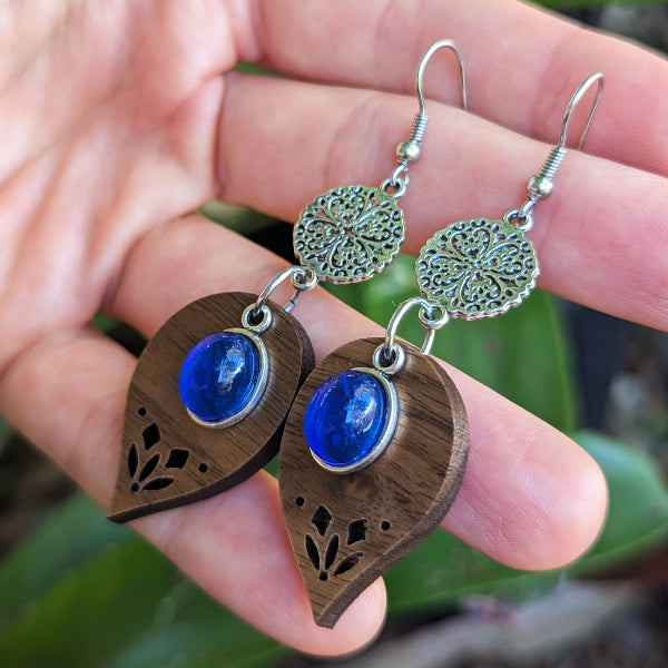 Wooden leaf shaped earrings with blue stones and silver accents held in a hand against a blurred natural background.
